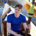 Mand sidder i en robåd og holder en åre. Baggrunden viser andre både i vandet og en bådebro. Han har en blå trøje på og ser alvorligt ud.