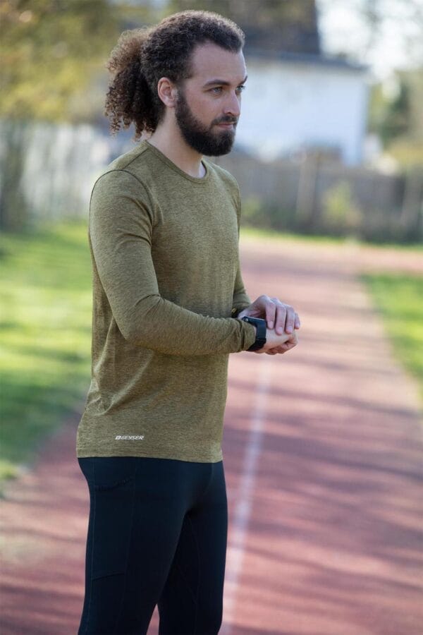 g21021_562_image_1 En mand med krøllet hår og skæg står på en løbebane iført en grøn trøje og sorte leggings; han justerer sit smartwatch. Trøjen har teksten "GEYSER".