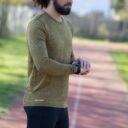 Mand iført en grøn trøje og sorte bukser, står og justerer sin smartwatch på en løbebane i udendørs omgivelser. Trøjen har "Geyser"-logoet nederst.