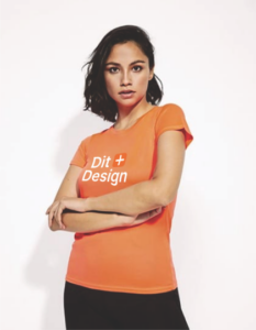 Person iført en orange T-shirt med teksten "Dit Design" trykt foran, står med armene over kors. Baggrunden er hvid og ensartet.