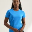 Person iført en blå t-shirt står stille med hænderne foldet foran sig i et hvidt rum.