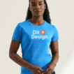 Person bærer en blå T-shirt med teksten "Dit Design" i et lyst rum. Personen står oprejst med hænderne let samlet foran sig.