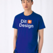 Person står iført en blå T-shirt med teksten "Dit Design" trykt på forsiden. Baggrunden er neutral med hvide gardiner.