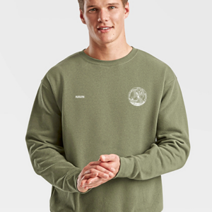 En ung mand iført en olivengrøn sweatshirt står med hænderne samlet. På trøjen står teksten "NAVN" og et rundet logo. Baggrunden er ensfarvet grå.