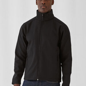 En person iført en sort jakke står i en neutral indendørs setting. Jakken er lukket, og personen har korte, mørke bukser på.