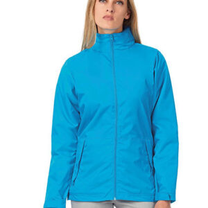 En person iført en blå jakke står mod en hvid baggrund. Jakken har en høj krave og lynlås, og personen har letbrunt langt hår.