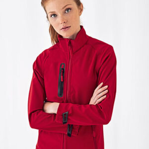 Person iført en rød jakke står med korslagte arme. Baggrunden er enkel og hvid, hvilket fremhæver tøjets farve og detaljer som lynlåse og syninger.