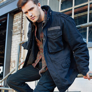 Person iført mørk jakke sidder på gelænder. Baggrund viser en murstensbygning med store vinduer. Personens blik er rettet mod kameraet. Ingen tekst til stede.