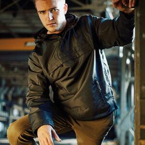 Jeg kan ikke genkende personer i billeder. En mand iført en sort jakke sidder på hug i et industrielt miljø med metalstrukturer og dæmpet belysning omkring ham.