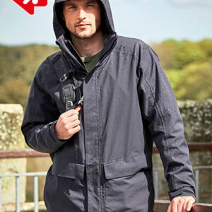 En person iført en mørk jakke med hætte står udendørs og holder jakken lukket. Baggrunden viser et grønt område med træer og en mur.