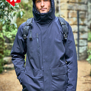 Mand iført mørk jakke og hætte bærer rygsæk, står på grussti omgivet af grønne planter og stenvægge. Rød logo grafisk element øverst til venstre.