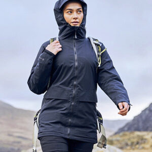 Person med rygsæk går i sort jakke med hætte i et bjergrigt landskab under en overskyet himmel.