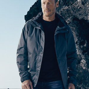 Mand står iført en mørk jakke og jeans ved siden af en klippeformation. Han kigger til siden, mens klipperne og en blå himmel dominerer baggrunden.