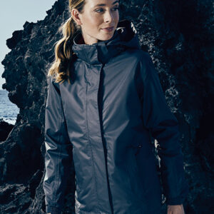 En person iført en mørk jakke står stille ved siden af en stenet baggrund i udendørs omgivelser. Der ses ingen tekst.