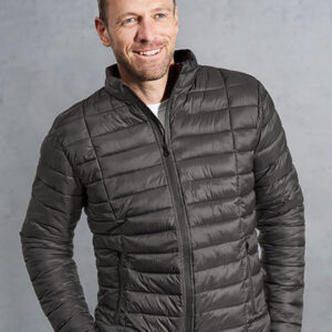 Mand i sort quiltet jakke smiler, med hænderne i lommerne, stående foran en neutral grå baggrund.