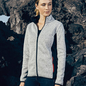 Person står iført en lysegrå jakke med lynlås, ser til siden foran en klippefuld baggrund. Stemningen er udendørs og rå.