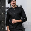 Kok iført sort kokkeuniform og sort baret smiler, mens han står i et køkkenmiljø med åbne hylder og køkkenredskaber i baggrunden.