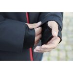 Hænder trækker en sort jakkeærme med tommelfingerhul op. Jakken har en rød lynlås og bæres i en udendørs gårdhave med brosten.