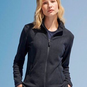 Person iført mørk fleecejakke, står afslappet med hænderne i lommerne, mod en lys blå baggrund. Personen har skulderlangt blondt hår.