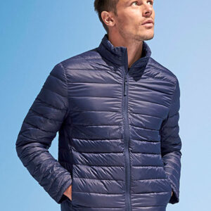 Mand iført en mørkeblå quiltet jakke kigger opad, med hænderne i lommerne. Baggrunden er en ensartet blå farve, der minder om en klar himmel.