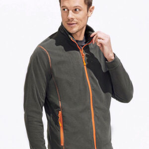 Mand klædt i en mørkegrøn fleecejakke med orange lynlåsdetaljer, står og kigger til siden mod en hvid baggrund. Han rører sin hage med højre hånd.