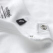 Hvid t-shirt ligger udfoldet med en sort mærkeseddel og to sølvknapper ved halsåbningen. Tekst: "Neutral Certified Responsibility" og "100% Organic Cotton".