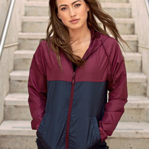Kvinde bærer en vinrød og mørkeblå jakke, står afslappet med hænderne i lommerne på en udendørs stentrappe, vind blæser let i hendes hår.