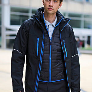 Mand i sort jakke med blå lynlåse står udendørs foran en moderne bygning. Han har også en hvid skjorte under jakke. Han ser fremad i en neutal stilling.