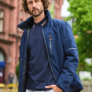 En person står afslappet og kigger fremad, iført en blå jakke og mørk trøje. Baggrunden viser en murstensbygning og grønne træer, hvilket giver en indbydende byatmosfære.