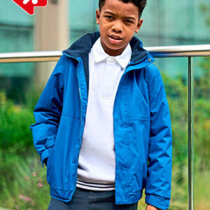 En dreng iført en blå jakke står afslappet med hænderne i lommerne. Han har en hvid skjorte og mørke bukser på. Baggrunden viser en glasbygning og grønne planter.