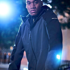 En person iført en sort jakke står udendørs om aftenen, omgivet af oplyste træer og svagt byggeri i baggrunden. Blåt og hvidt lys skinner i nærheden.