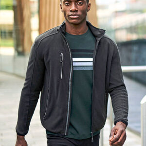 Person går på en udendørs gangbro, iført en sort jakke og en grøn t-shirt med hvide og grå striber. Baggrunden viser moderne bygninger og svage træstrukturer.