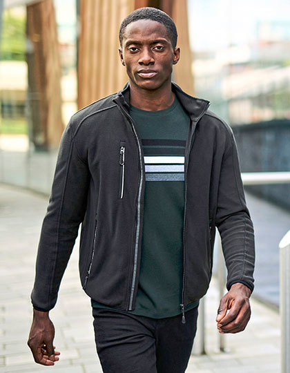 Person går på en udendørs gangbro, iført en sort jakke og en grøn t-shirt med hvide og grå striber. Baggrunden viser moderne bygninger og svage træstrukturer.