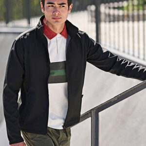 En person står med én hånd på et metalrækværk. Personen bærer en sort jakke over en hvid trøje med farvede striber og har kort mørkt hår. Baggrunden er udendørs.