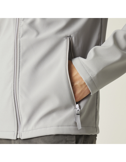 RG762_6_Detail En person iført en lys jakke stikker deres hånd ind i en sidelomme. Personen står i en neutral, minimalistisk indendørs omgivelser.