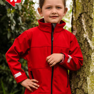 Et barn iført en rød jakke står smilende ved et træ i en grøn, udendørs omgivelser. Ingen tekst er synlig i billedet.