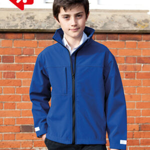 En person iført en blå jakke står stille foran en væg lavet af røde mursten. Vedkommende har hænderne i lommerne og ser direkte fremad.