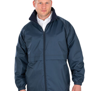 Mand står iført en blå jakke med lynlås og krave, hænderne afslappet ved siderne, i et neutralt, hvidt baggrundsmiljø. Han bærer en hvid skjorte under jakken.