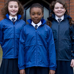 Tre skolepiger står sammen iført ens blå jakker og slips, smilende, foran en murstensbygning med en buet sort dør i baggrunden.