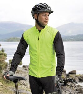 En person iført en gul refleksvest og sort cykelhjelm står med en cykel ved en stenstrækning. I baggrunden ses bjerge og en sø i et naturlandskab.