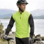 En person iført en gul refleksvest og sort cykelhjelm står med en cykel ved en stenstrækning. I baggrunden ses bjerge og en sø i et naturlandskab.