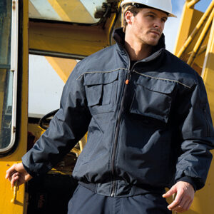 Mand med sikkerhedshjelm og mørk jakke står ved en stor, gul maskine, som kunne være en byggekran. Han kigger opmærksomt fremad, omgivet af metalstrukturer i dagslys.