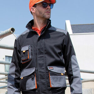 Mand iført sort og grå arbejdstøj og orange kasket kigger fremad. Han står ved nogle metalrør med en moderne bygning i baggrunden under en blå himmel.