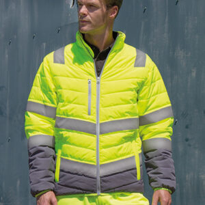 Mand står iført en neongul jakke med reflekterende striber, hænderne i lommerne, foran en metalvæg. Han har kort hår og kigger til siden.