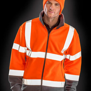 Person iført orange refleksjakke og hue, står med hænderne i lommerne, isoleret mod en mørk baggrund. Der er intet synligt tekst.