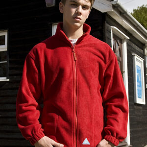 En person iført en rød jakke står stille foran en mørk bygning med hvidt rammeværk. Sollys skaber skygger på væggen bagved.