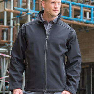 Mand i sort jakke står afslappet på byggeplads med stilladser omkring sig og bærer grå skjorte og bukser.