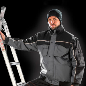 En person iklædt en grå jakke og sort hue hviler sin hånd på en stige, med en sort baggrund. Personen ser direkte fremad.