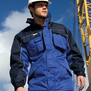 Mand i blå arbejdsjakke kigger fremad; han bærer en hvid hjelm, står foran en kran under en blå himmel med skyer.