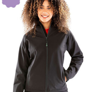 En person med krøllet hår bærer en sort jakke og står smilende. Personen har en rød trøje under jakken, og baggrunden er hvid med en lilla detalje øverst.